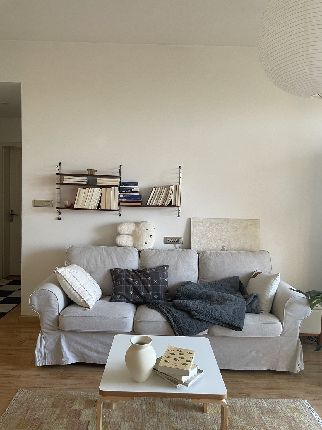 Grey slipcovered sofa with cushions and a dark throw, a white coffee table holding a cream Crock Style Vase and books, on a faded patchwork rug