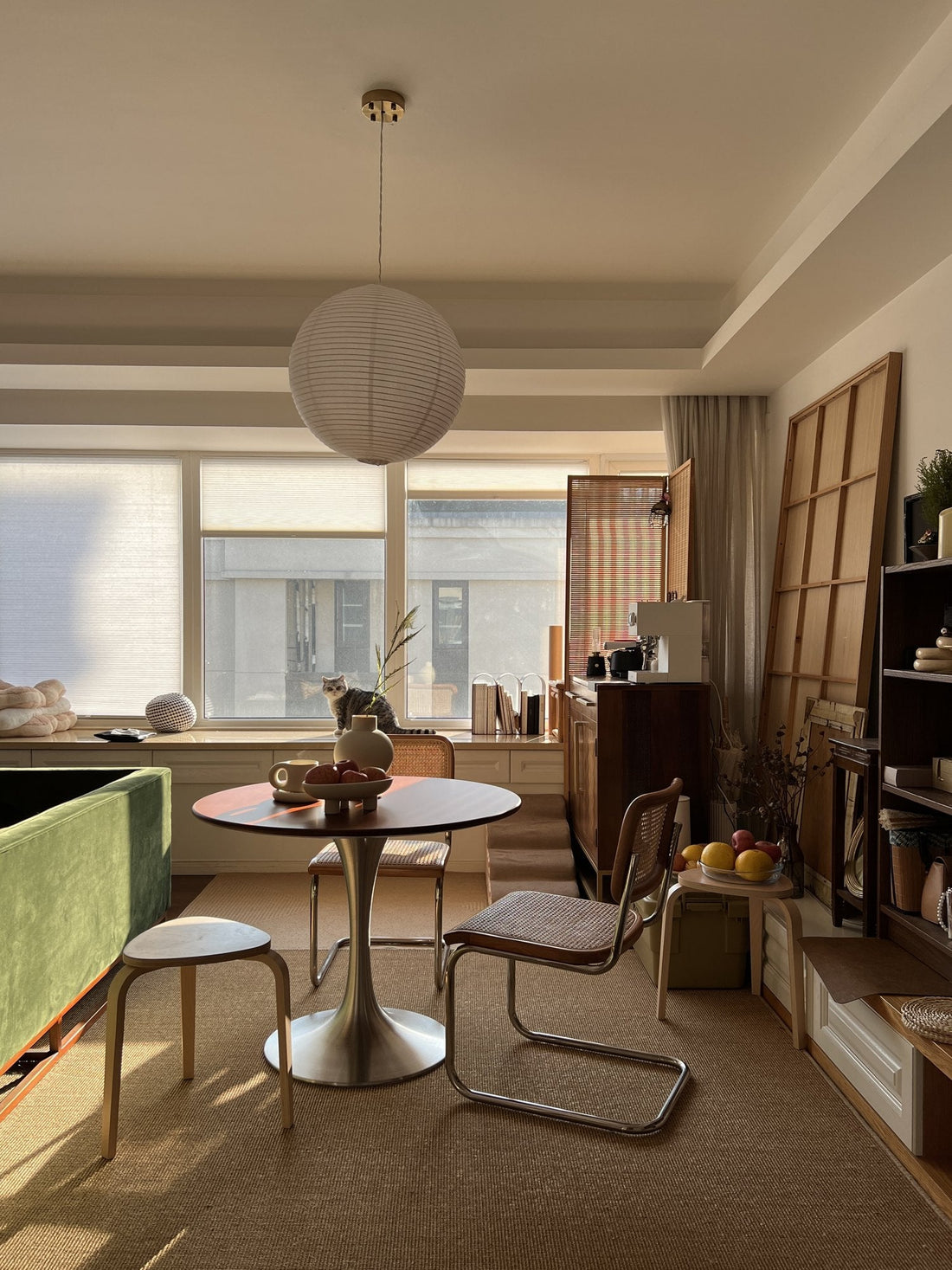 Sunlit room with a round wooden dining table on a brushed metal base, cane chairs, a paper lantern and a green velvet sofa