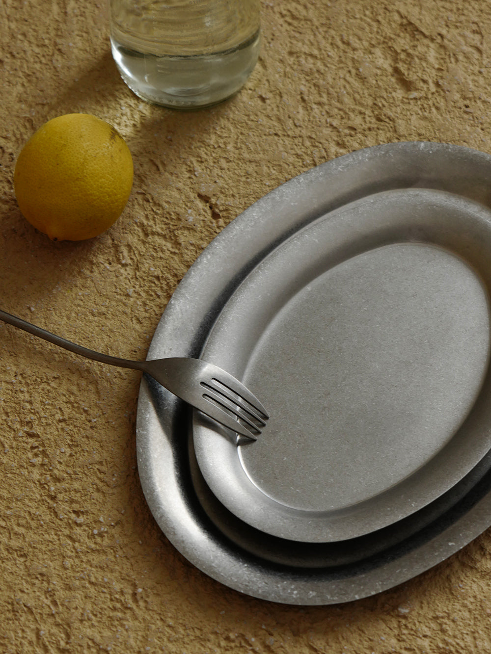 Two Ova oval stainless steel plates nested on a textured table beside a fork and lemon