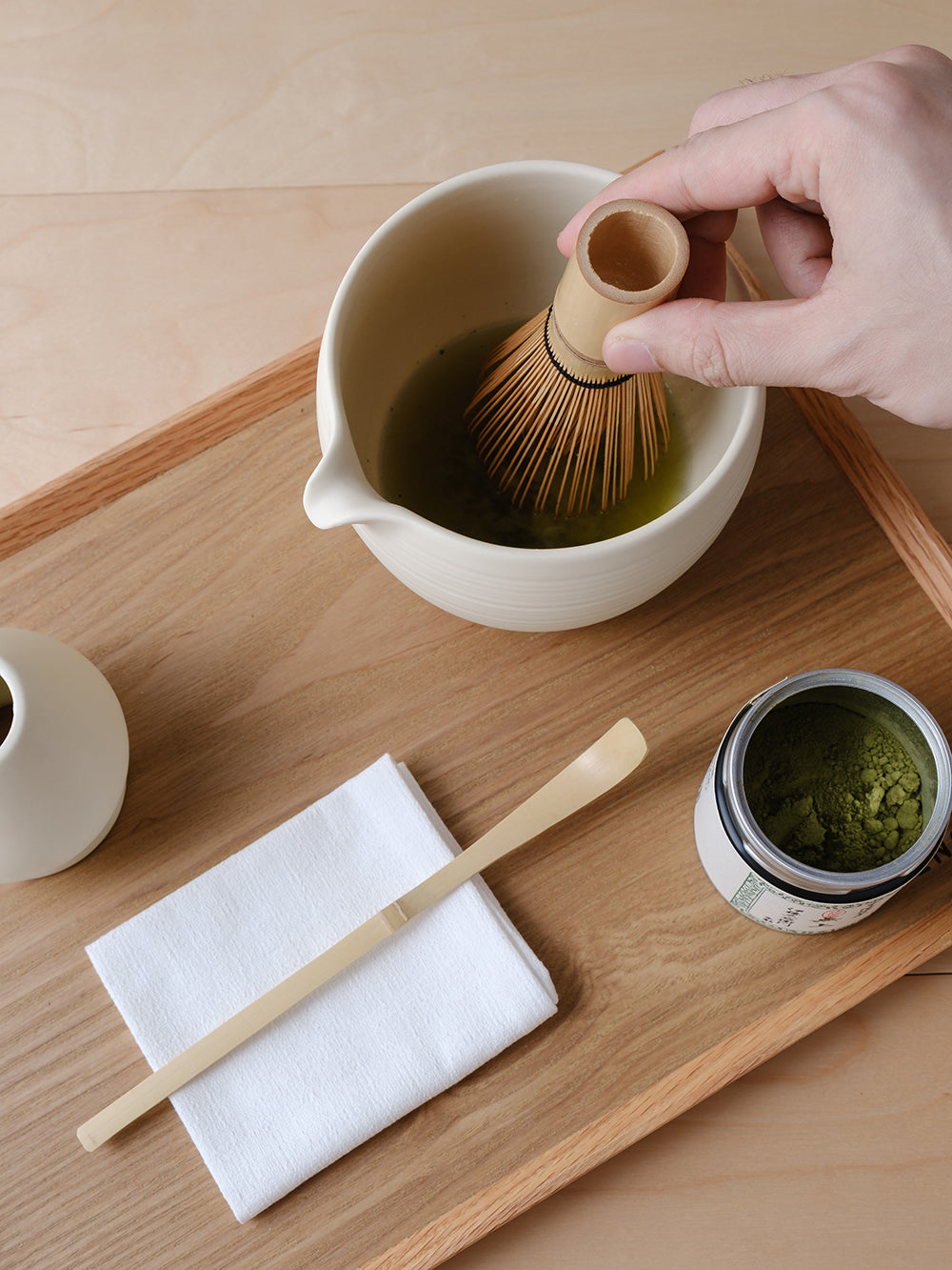 A bamboo whisk being lifted from a cream ceramic matcha bowl with a spout on a wooden tray