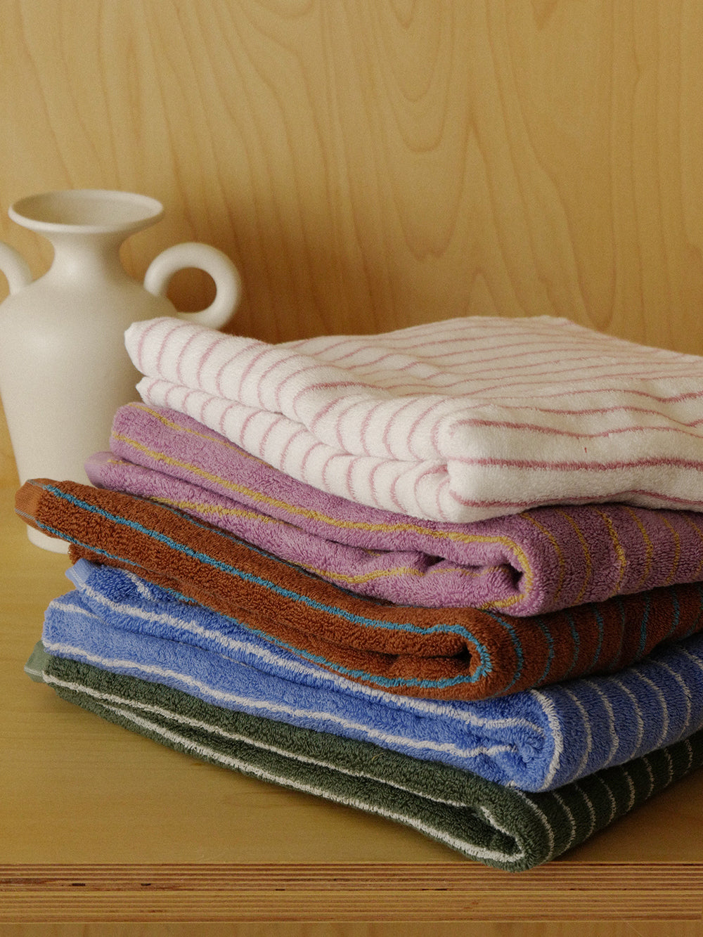 Striped cotton towels in pink, purple, caramel, blue and green folded in a stack on a wooden shelf beside a cream ceramic vase