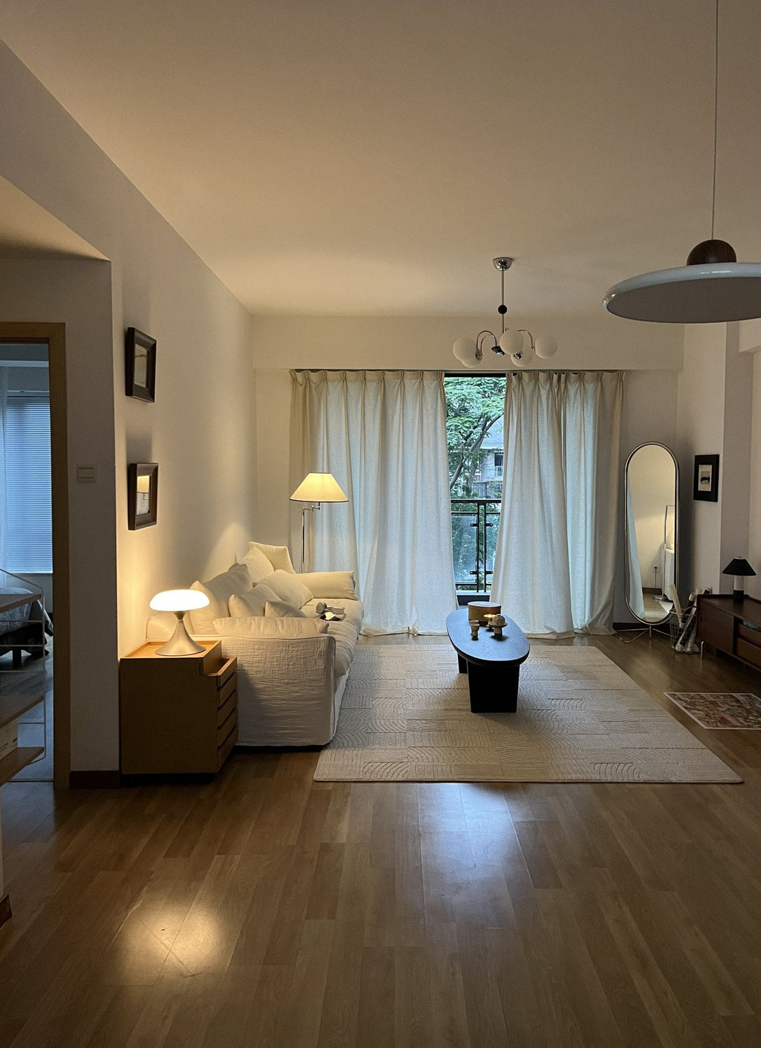 Living room at dusk with a cream sofa, black oval coffee table, pale rug and floor lamp in front of linen curtains