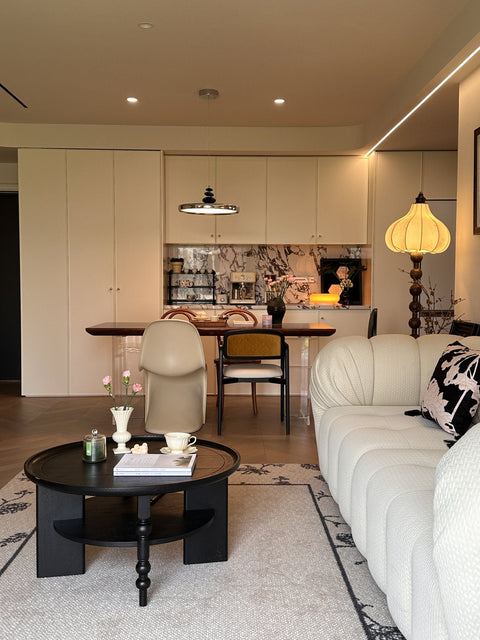 Living room in a French mid-century style with a cream channel-tufted sofa, a round black coffee table, a pumpkin floor lamp and the dining area beyond