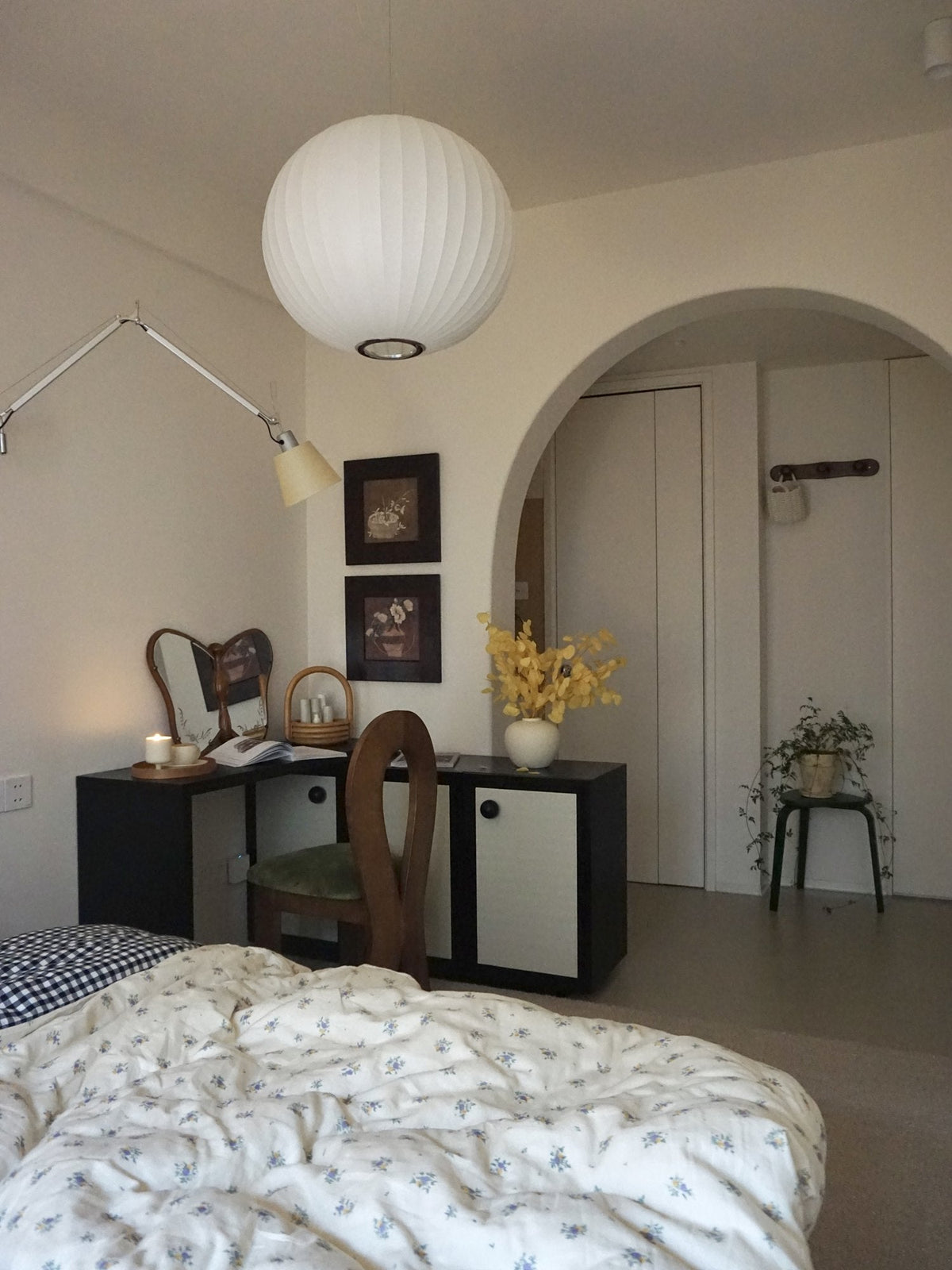 Bedroom with a black desk and white cupboards, wooden chair, yellow flowers in a cream vase and a paper pendant above an arched doorway