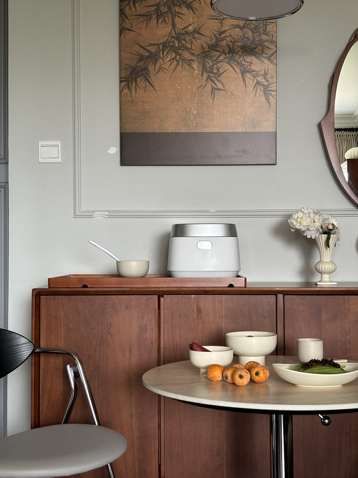 Round stone-topped table with footed cream bowls, a dish of loquats and a plate of greens in front of a cherry-wood sideboard