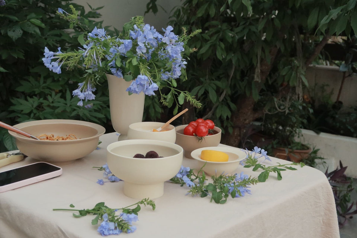 Garden table with a cream cloth, footed cream bowls of bayberries and tomatoes, a wide-rim bowl of pasta and blue plumbago in a cream vase