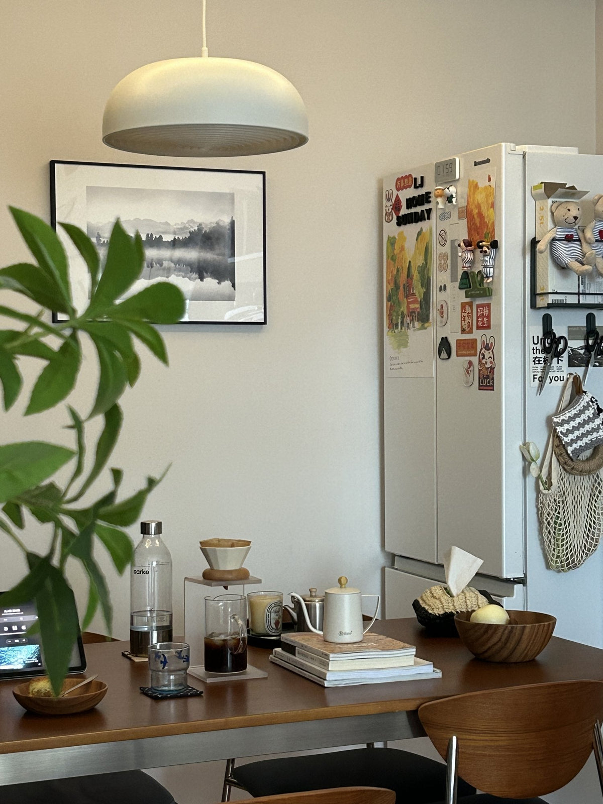 Dining table by the kitchen with a pour-over coffee stand, a kettle, books and a bowl of fruit, under a white dome pendant
