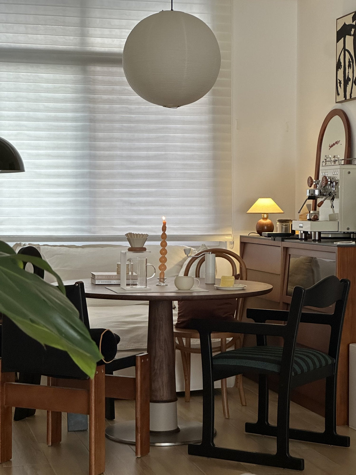 Dining corner with a round wooden table, black and wood chairs, a paper globe pendant and a sideboard with a coffee machine under the window