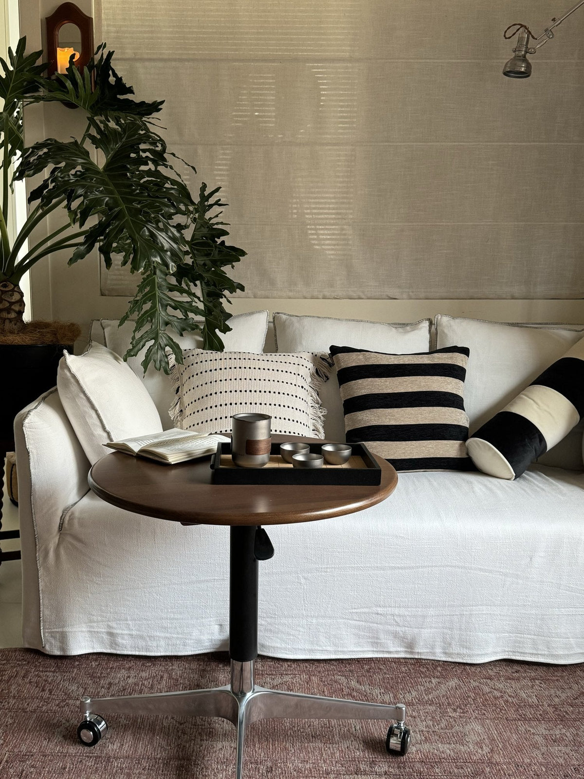 White linen sofa with striped cushions and a large split-leaf plant, with a round wooden side table on castors holding a tea set on a tray