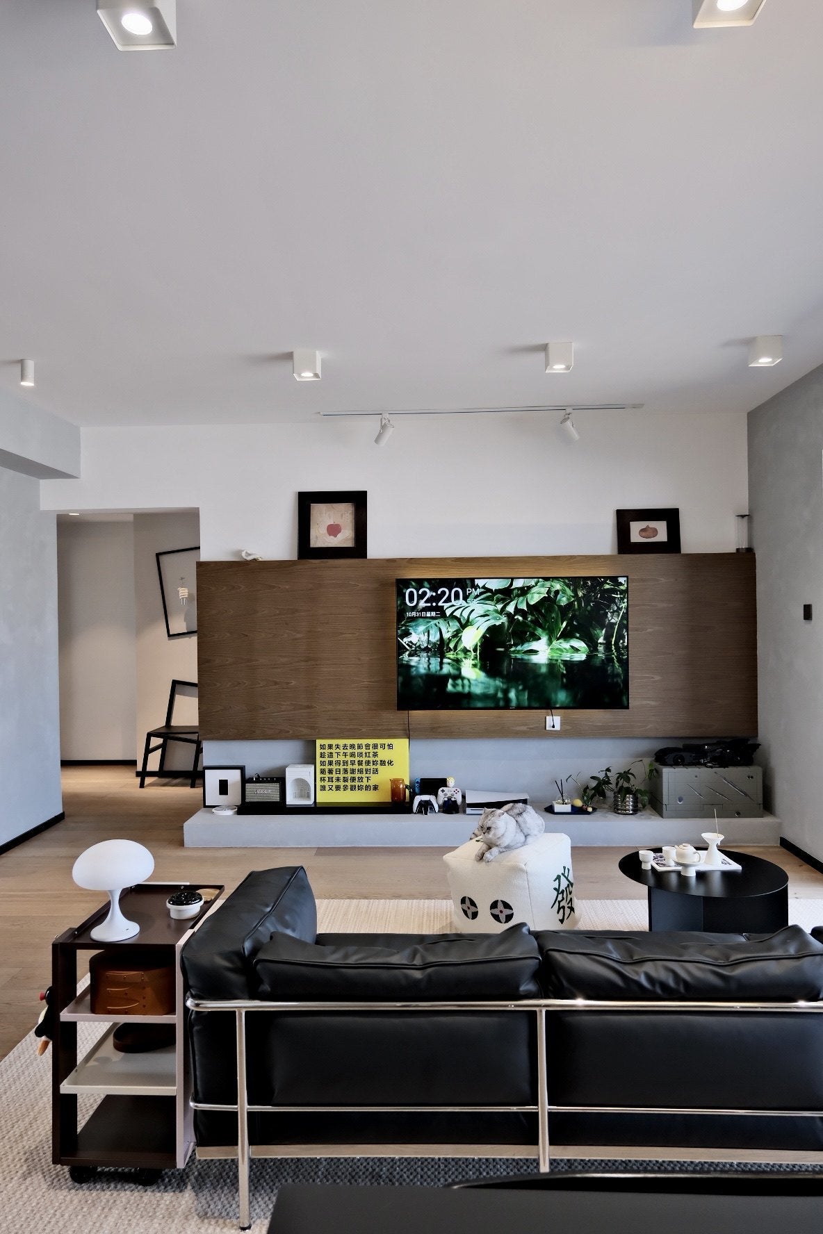 Open living room with a black leather sofa, a round black coffee table, a white cube pouf and a wood-panelled television wall