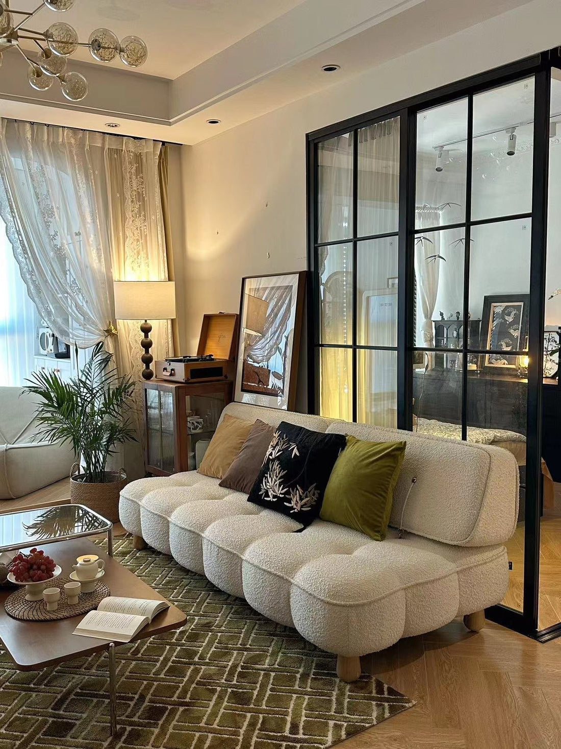 Living room with a cream bouclé sofa, embroidered cushions, a green patterned rug and a walnut coffee table, beside a black-framed glass partition