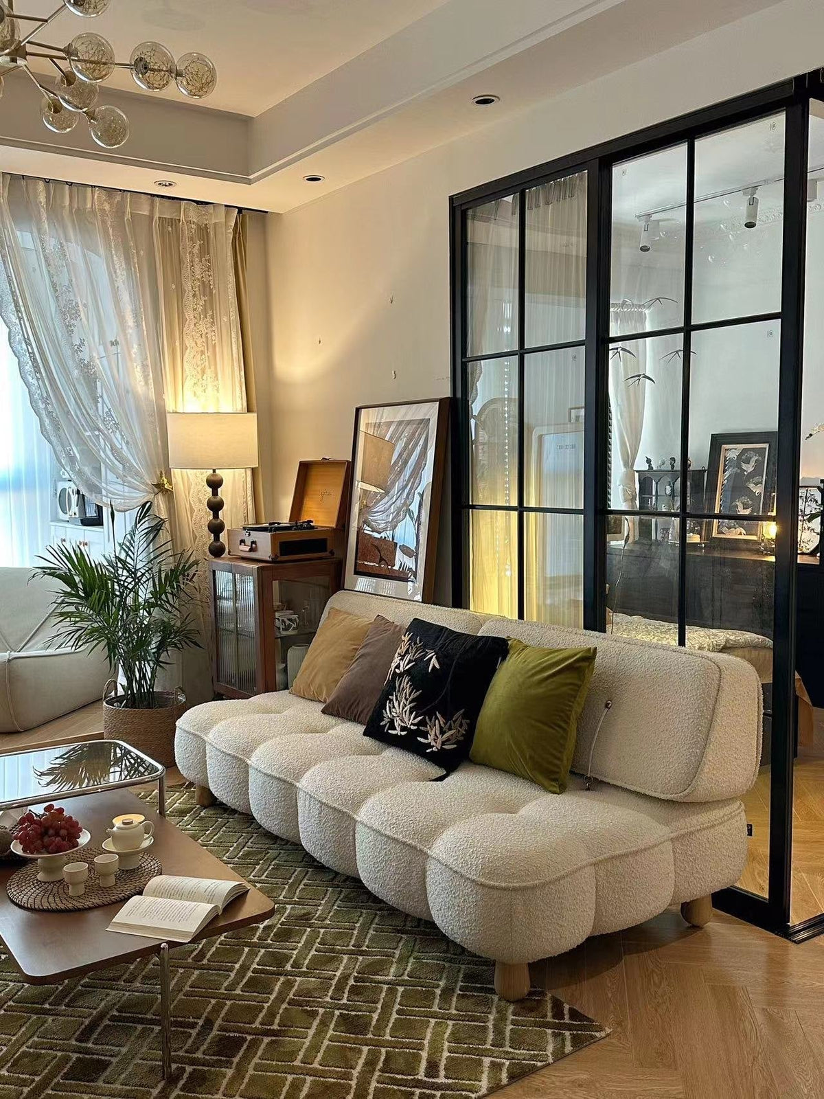 Living room with a cream bouclé sofa, embroidered cushions, a green patterned rug and a walnut coffee table, beside a black-framed glass partition