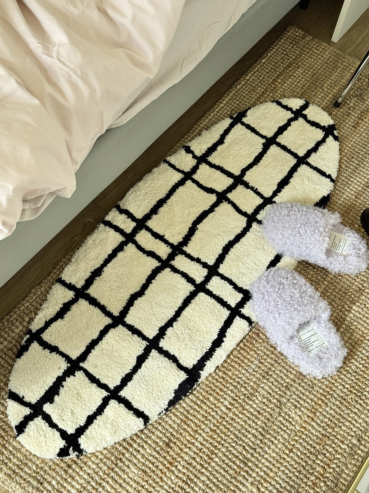Oval cream and black grid rug beside a bed and slippers